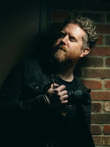 Man with curly hair and beard holding a camera near a brick wall.