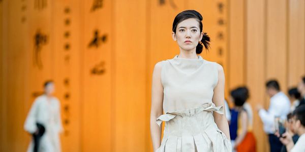 Model walking on runway in a cream dress with an orange background.
