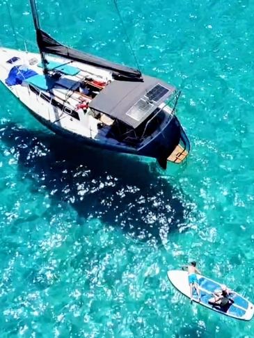Sailboat anchored in crystal-clear turquoise water with two people on a paddleboard nearby.