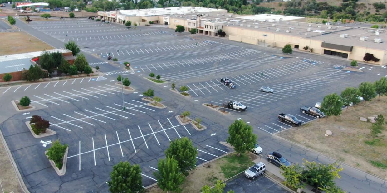 A nearly empty large parking lot with a few cars and a shopping center in the background.