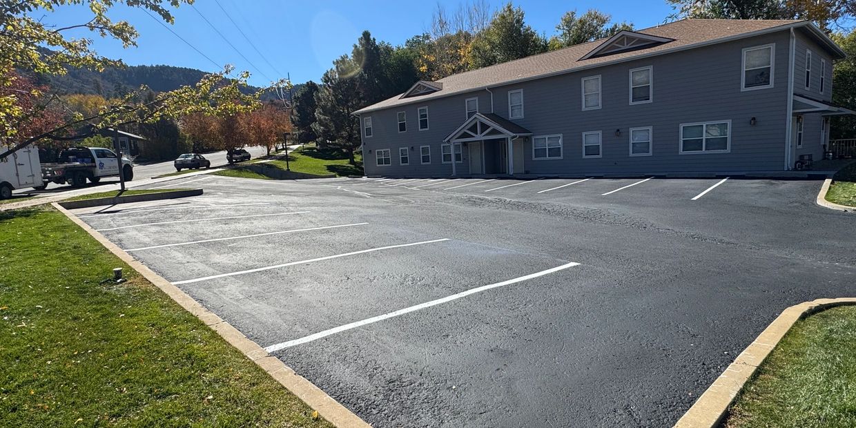 Empty parking lot beside a two-story gray building on a sunny day.