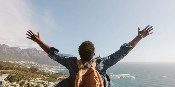 A person looking out to the sea, throwing their arms in the air in joy of achievement.