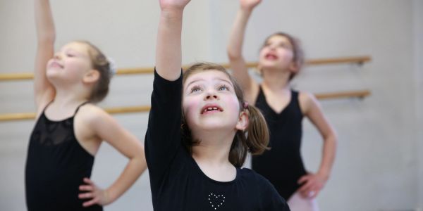 Three young girls in black dance outfits reaching upward during ballet class.