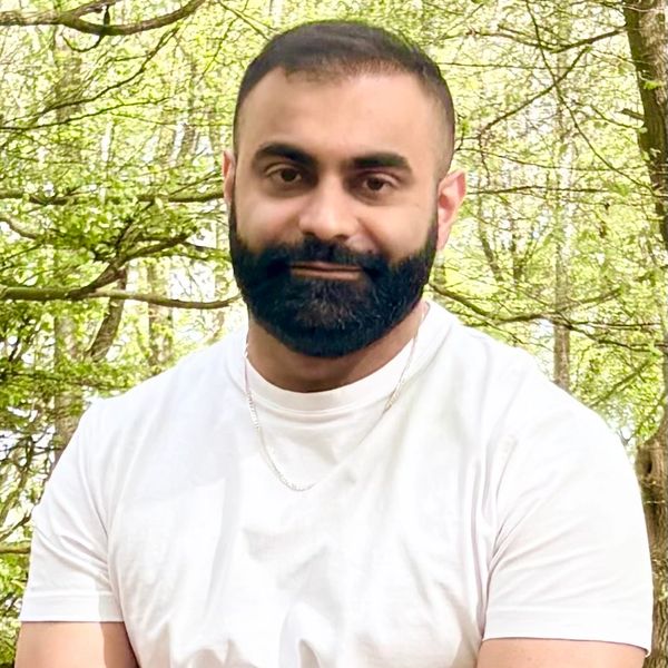 Man with a beard wearing a white t-shirt outdoors with trees in the background.