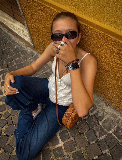 Young woman sitting on cobblestone street against a yellow wall, wearing sunglasses and casual clothes.