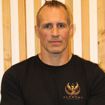 Man wearing a black Alchemy shirt against a wooden backdrop.