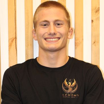 Young man smiling in a black Alchemy t-shirt against a wooden background.