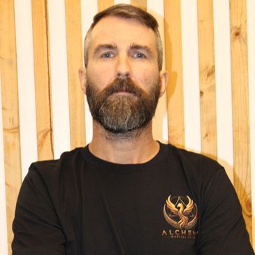 Bearded man in black Alchemy shirt stands against wooden slats.
