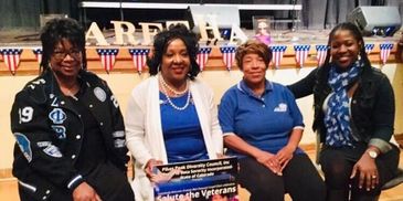 Four women seated together at a veterans event, smiling and holding a sign.