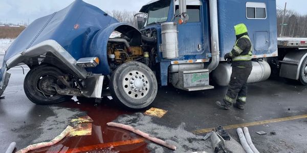 Damaged blue semi-truck with a person inspecting spilled fluid on the ground.