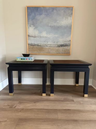 Two black side tables under a framed beach painting.