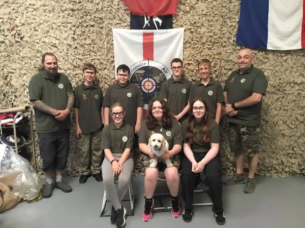 Group photo of air rifle club members in uniform posing indoors with a dog.