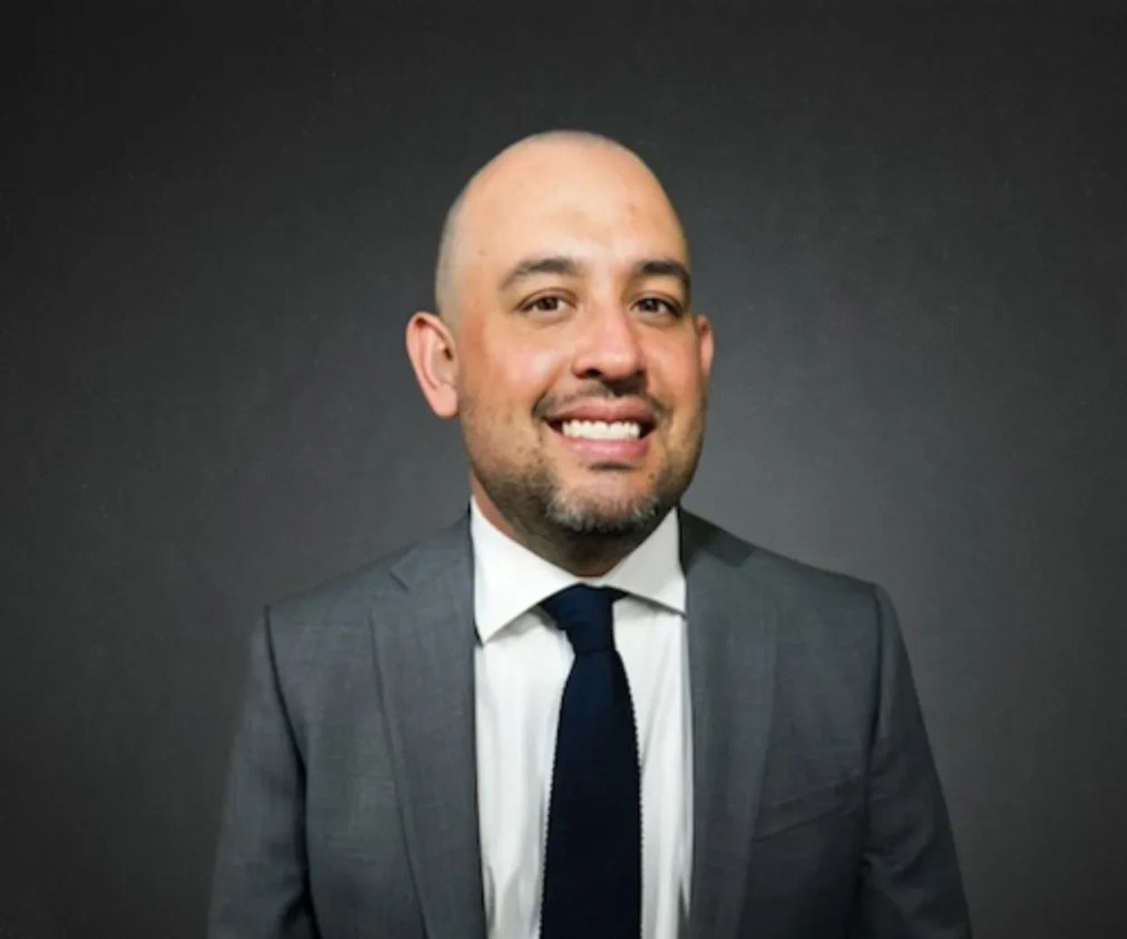 Smiling man in a gray suit and navy tie against a dark background.