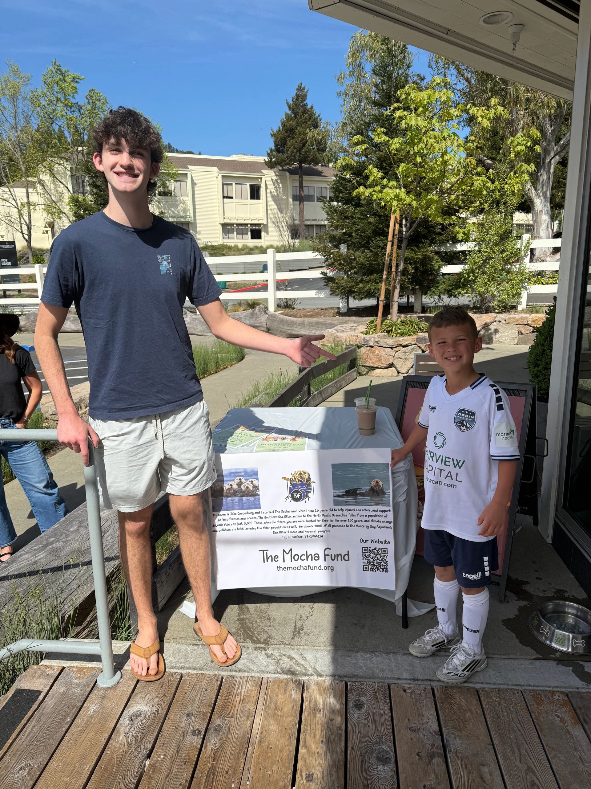 Two boys standing by a table promoting The Mocha Fund on a sunny day.