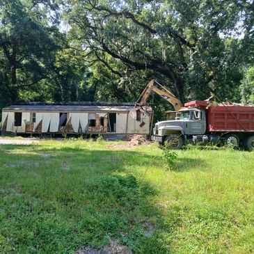 Excavator demolishing an old trailer with a dump truck nearby.