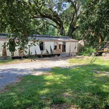 Old, damaged mobile home surrounded by trees and a construction vehicle.