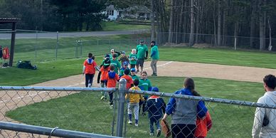 Children and adults walking on a baseball field with trees in the background.