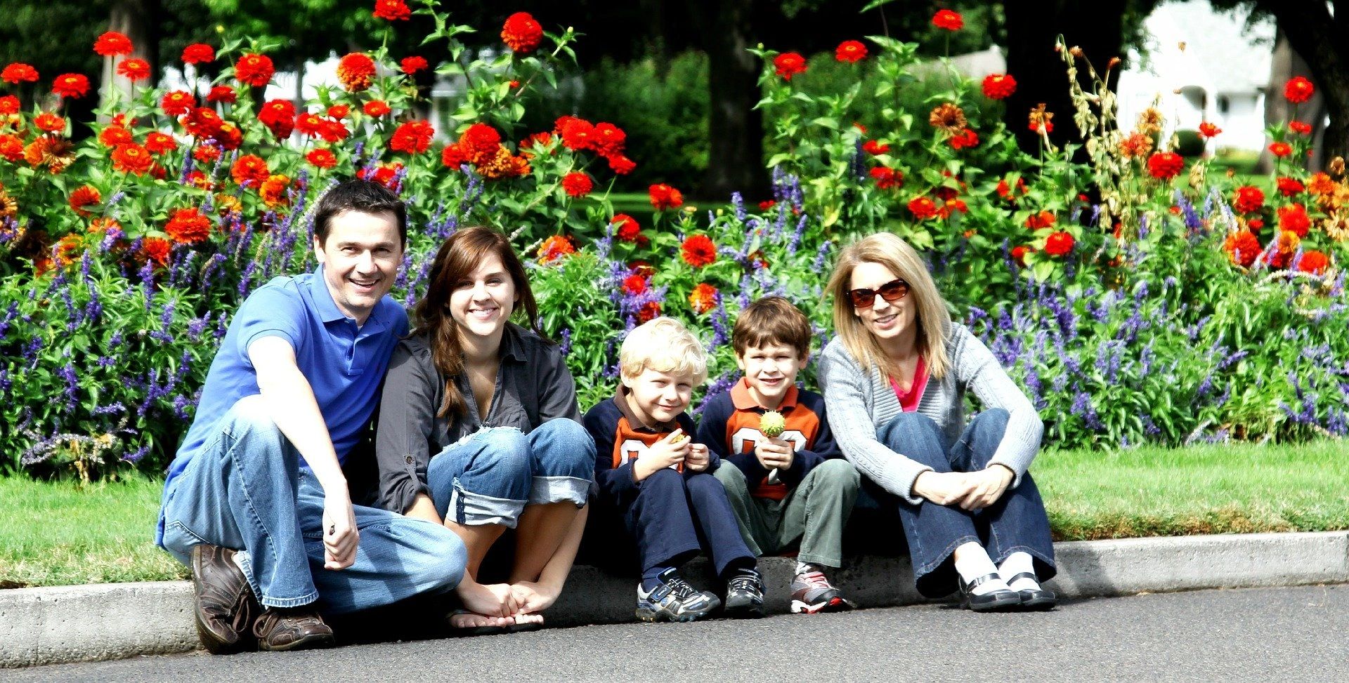 Family of five sitting on a curb in front of red roses and purple flowers.