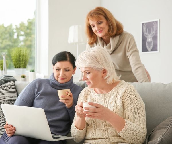 Three women gathered around a laptop, enjoying coffee together at home.