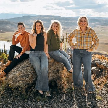 Four women posing outdoors on a sunny day with mountains in the background.