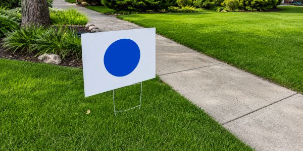 White yard sign with a large blue dot on green grass near sidewalk.