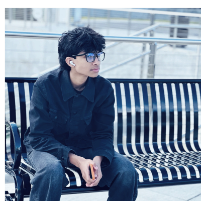 Young man with glasses and earbuds sitting thoughtfully on a bench.