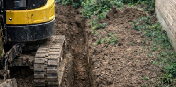 Mini excavator digging a narrow trench beside a building.