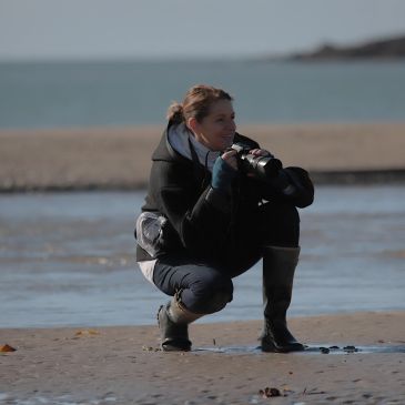 Woman crouching on beach with binoculars, looking out to sea.