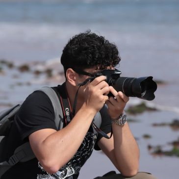 A man with curly hair taking a photo with a Canon camera near water.
