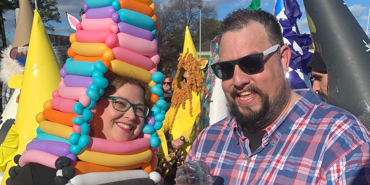 A woman in a colorful balloon hat smiles next to a man in sunglasses and a plaid shirt.