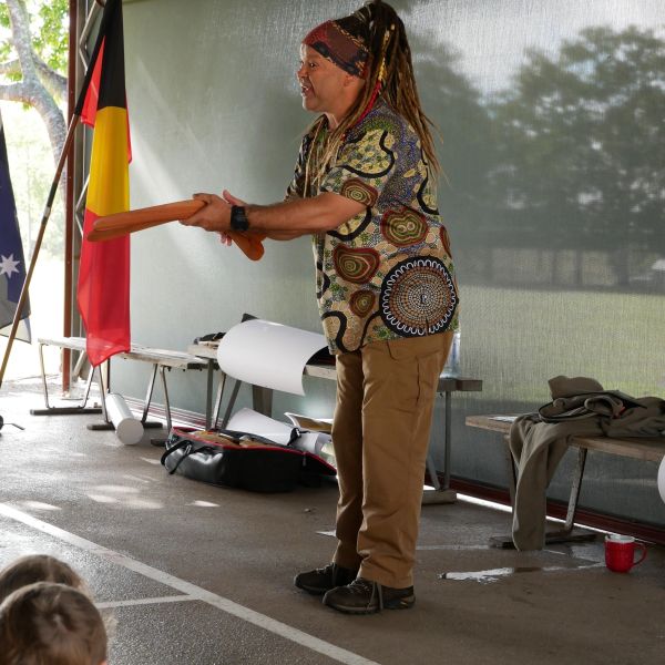 Man with dreadlocks holding a wooden instrument, performing indoors near flags.