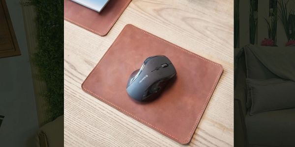 A black computer mouse on a brown leather mouse pad on a wooden desk.
