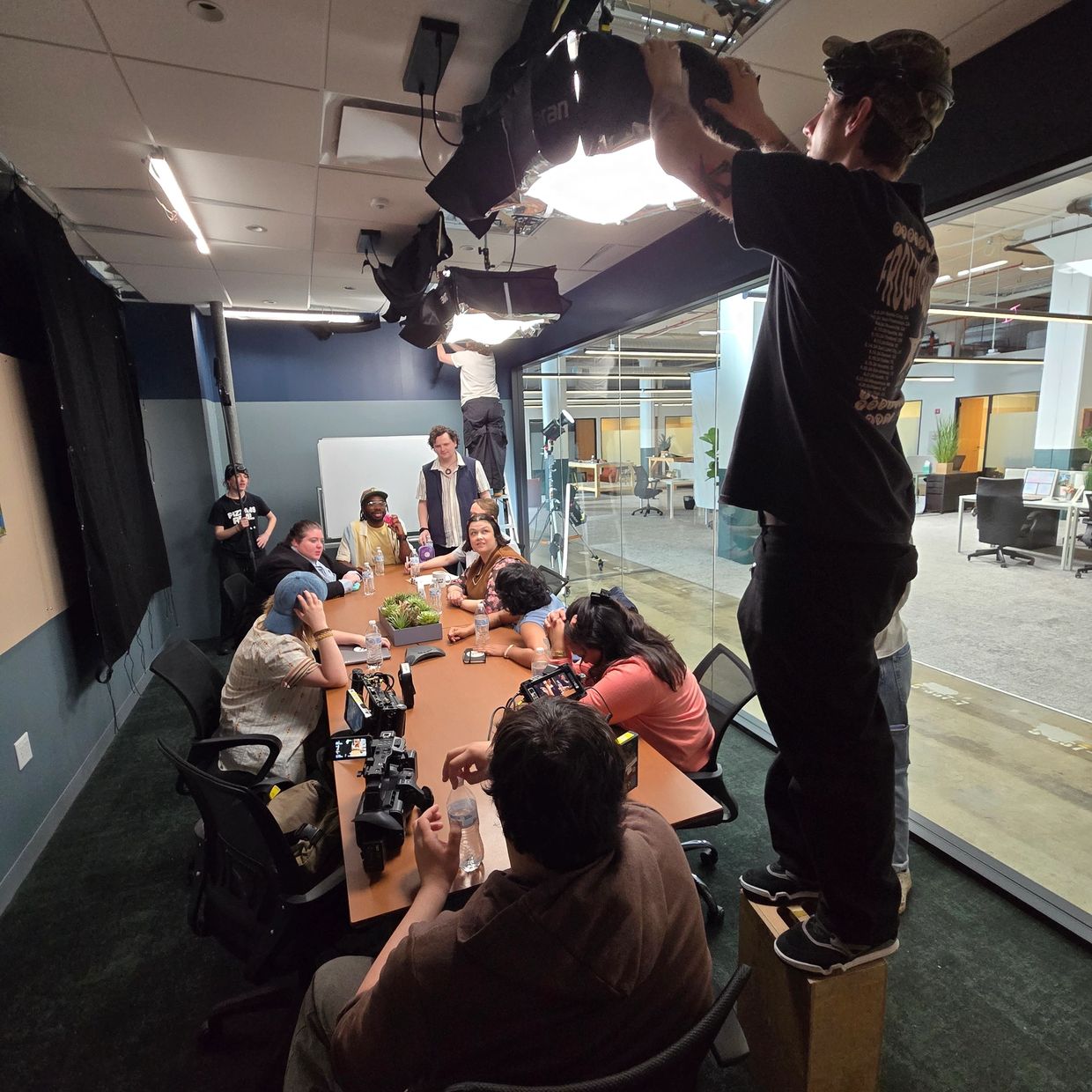 Film crew setting up lighting and cameras around a meeting table with people.