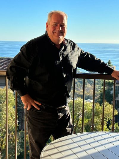 Man standing on balcony overlooking ocean and houses at sunset.