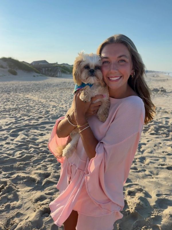 Smiling woman holding a small dog on the beach at sunset.