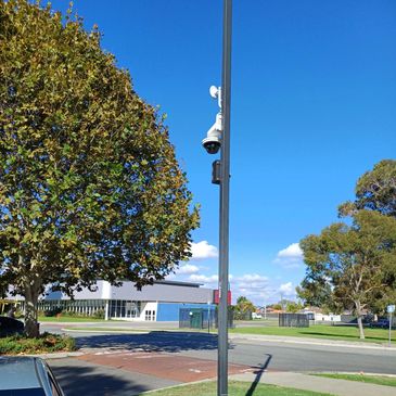 A streetlight pole with security cameras under a clear blue sky near a tree and buildings.