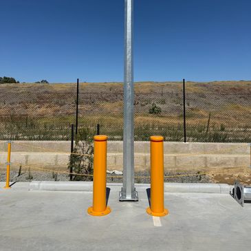 Two bright orange bollards protect a metal pole in a parking area under clear blue sky.