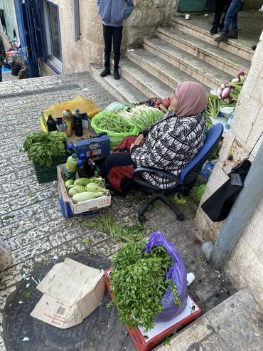A woman selling fresh vegetables at a street market on stone pavement.