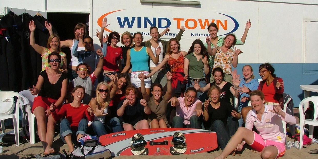 Group of happy women posing in front of a kitesurfing shop with a kiteboard on sand.