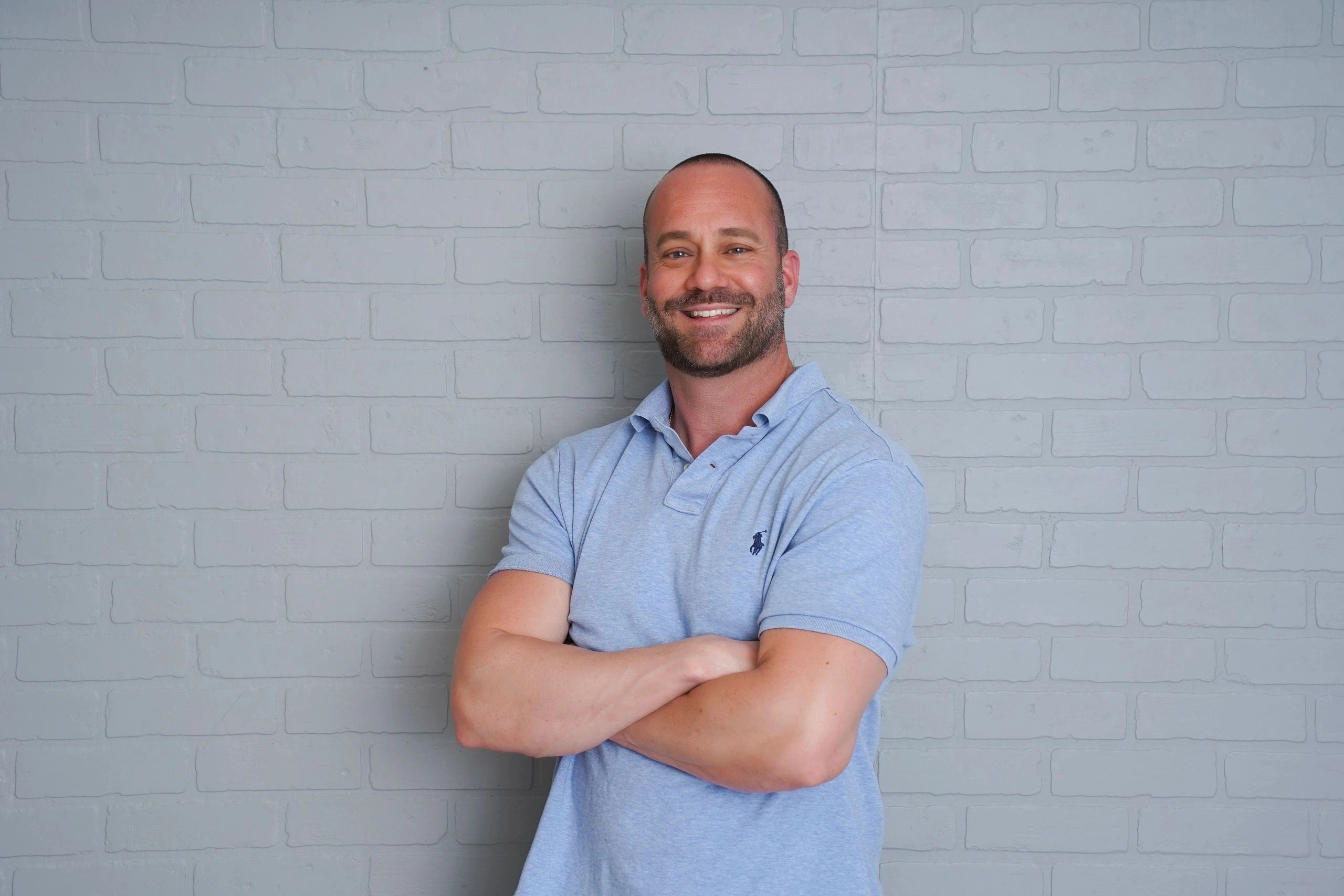 Smiling man with crossed arms in blue polo shirt against gray brick wall.