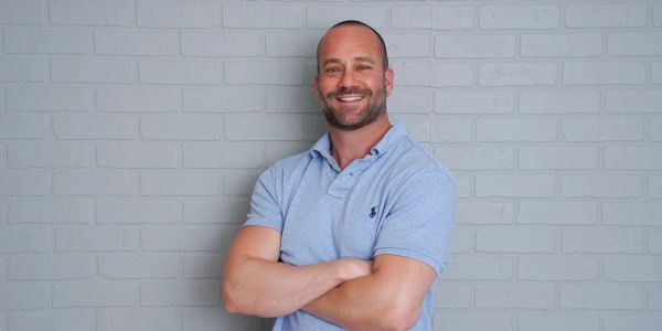 Smiling man with crossed arms stands against a gray brick wall.