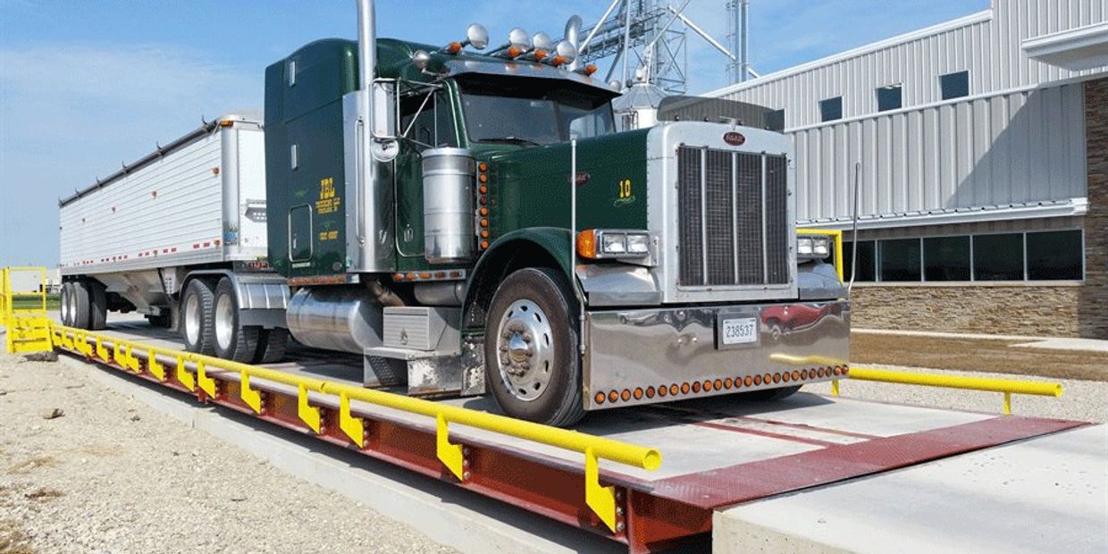 A green semi-truck on a weighbridge outside a warehouse.