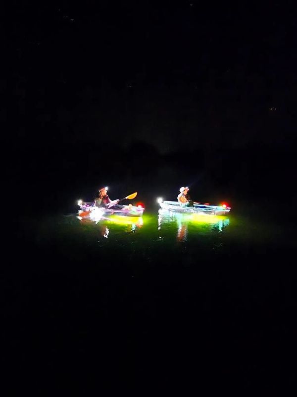 Two kayakers illuminated by bright lights paddle at night on calm water.