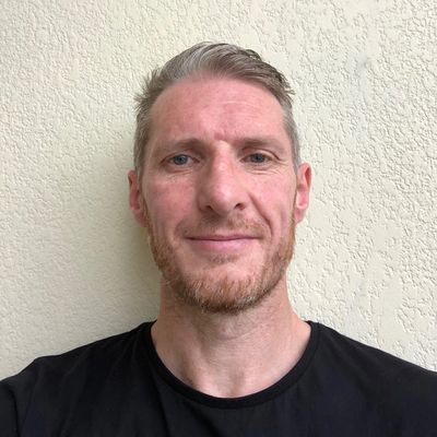 Smiling man with short hair and beard against a textured wall.