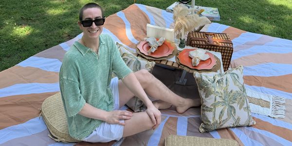 Person enjoying a stylish picnic on a patterned blanket with cushions and tableware.