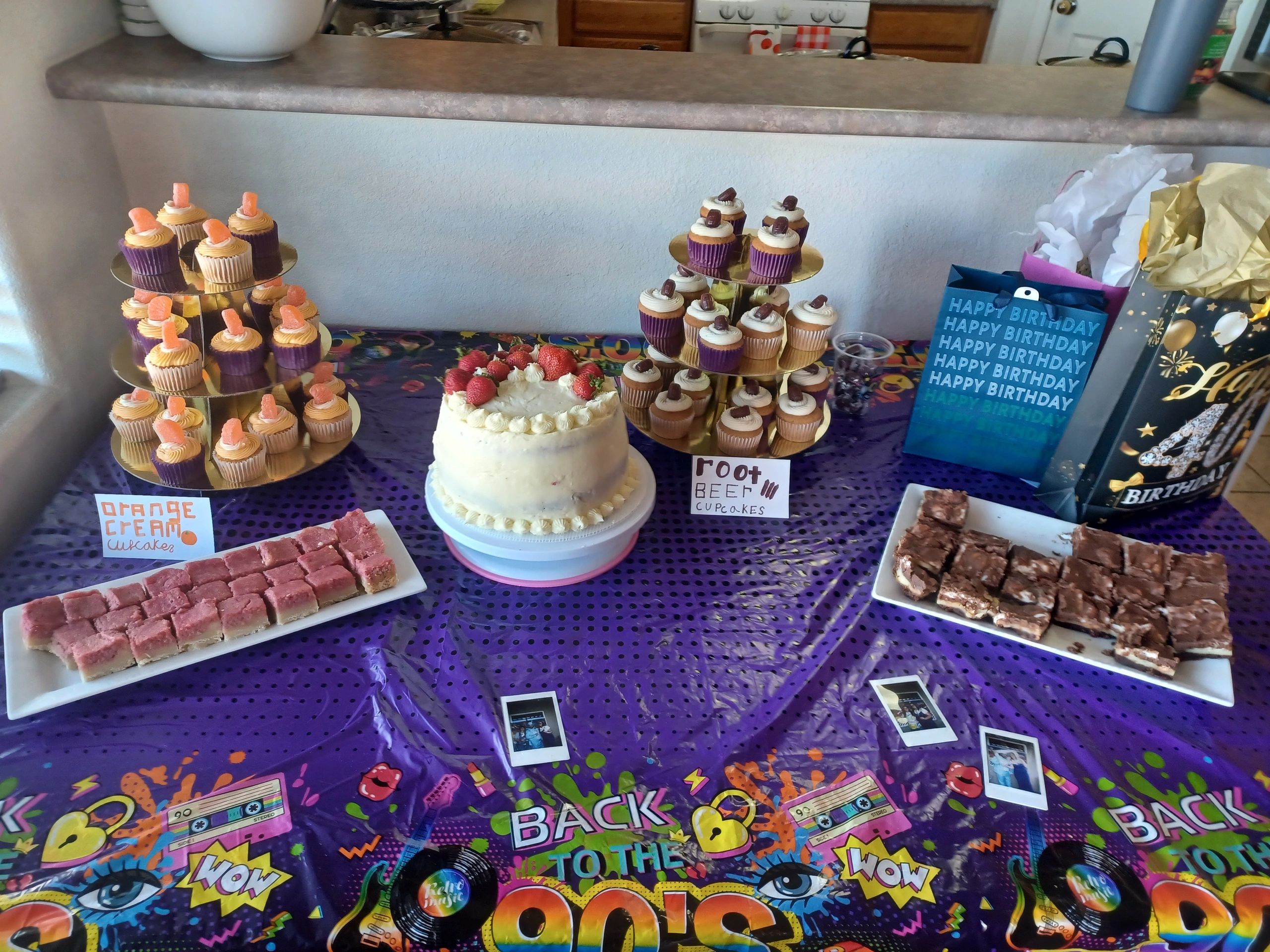 Birthday dessert table with cupcakes, cake, and brownies on a colorful 90s-themed tablecloth.