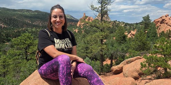 Woman sitting on a rock with a mountainous forest backdrop under a partly cloudy sky.