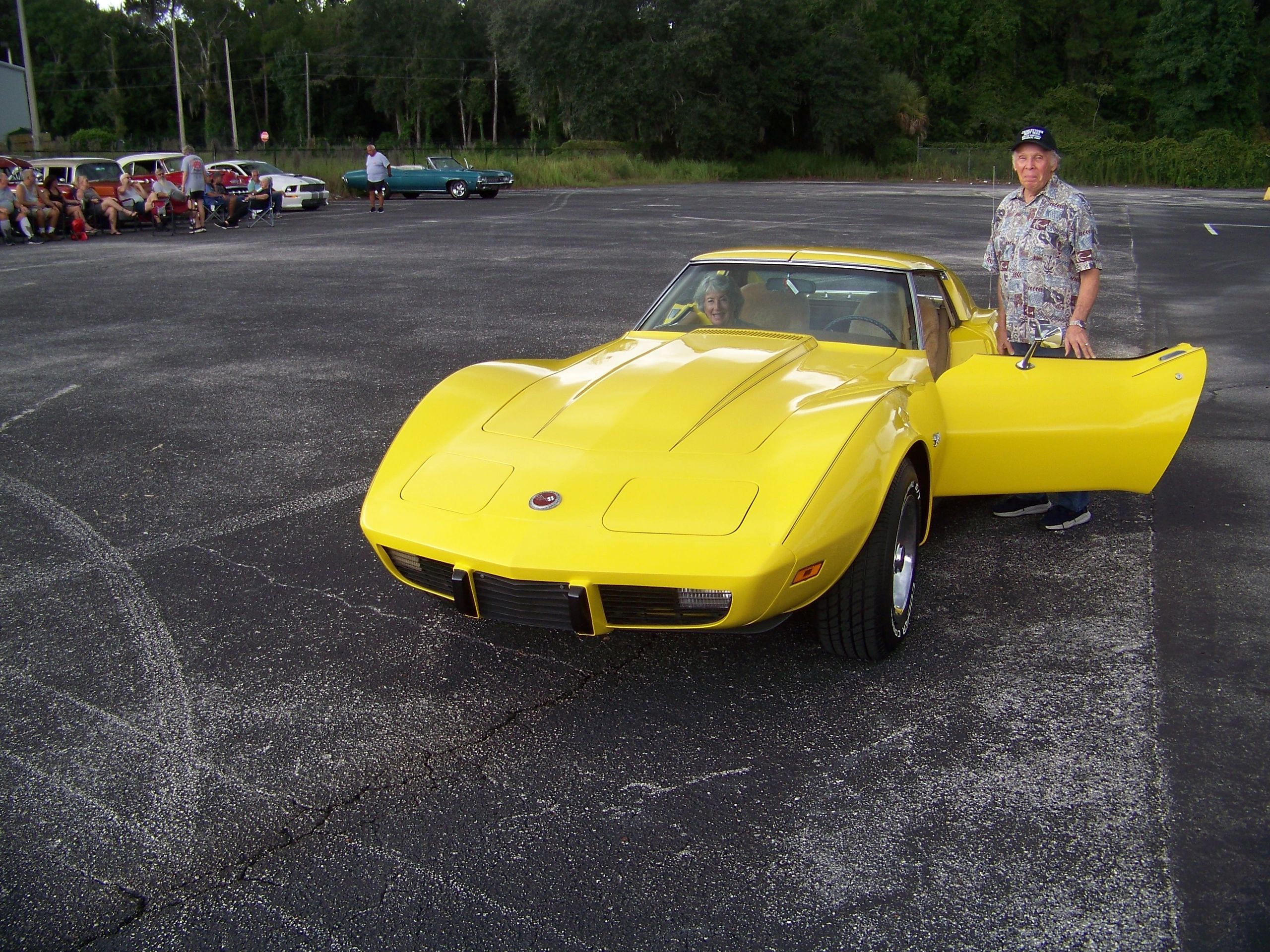 Man stands by a yellow classic car with woman inside and people sitting near vintage cars in background.