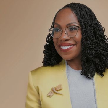 Smiling woman with glasses and curly hair wearing a yellow blazer and a bee brooch.