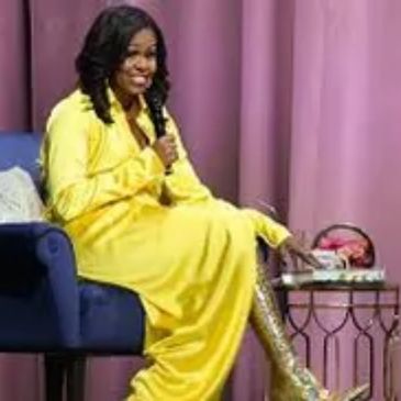A woman in a bright yellow dress sitting on a blue chair, speaking into a microphone.